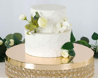 May include: Gold cake stand with a crystal beaded trim. A two-tiered white cake with white flowers is on the stand.