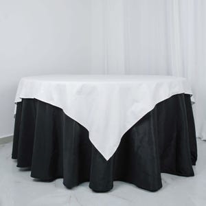 May include: A round table covered with a two-tone tablecloth. The top layer is white, and the bottom layer is black. The white layer is square-shaped, and the black layer drapes to the floor.