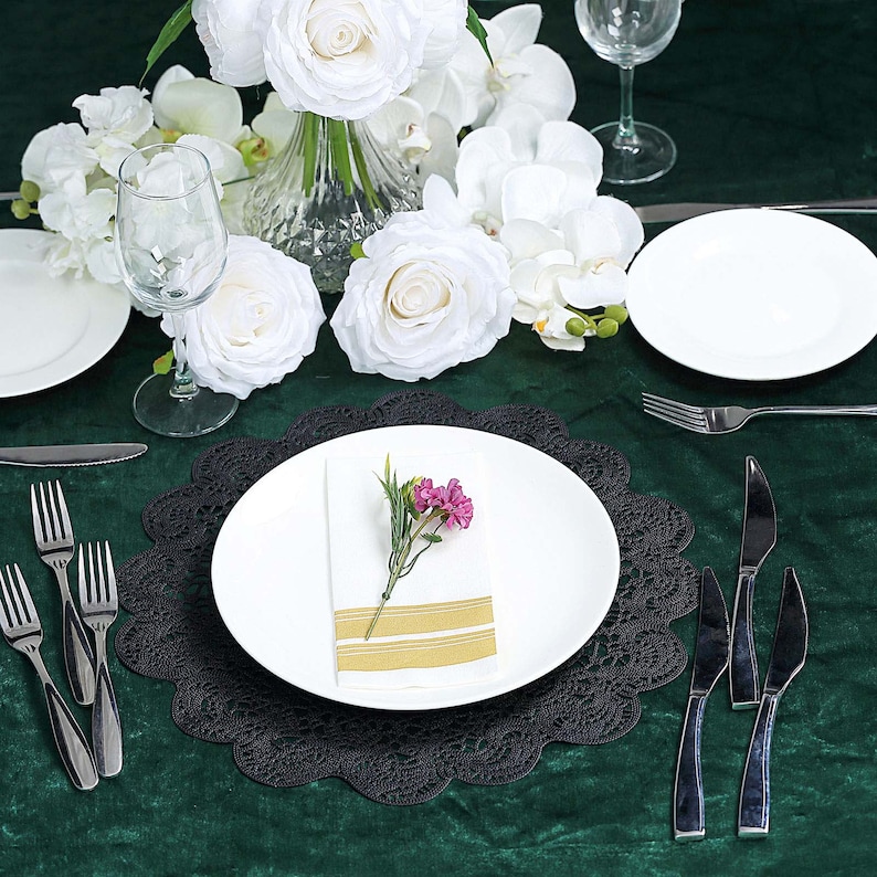 Puede incluir: Un plato de servicio de encaje negro con un plato de cena blanco en la parte superior. Una servilleta blanca con una flor rosa se coloca en el plato. El plato de servicio est&aacute; sobre un mantel verde.