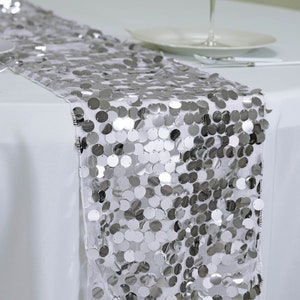 May include: Silver sequin table runner with round sequins. The runner is on a white tablecloth.