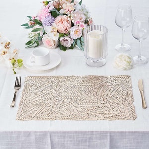 May include: A beige placemat with a delicate leaf pattern design. The placemat is set on a white tablecloth with a white cup and saucer, a glass candle holder, and a bouquet of pink, white, and purple flowers.
