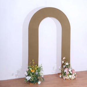 May include: A large, arched wedding backdrop in a neutral tan color. The arch is decorated with floral arrangements at the base, featuring white, yellow, and pink flowers. The backdrop stands on a wooden floor against a white wall.