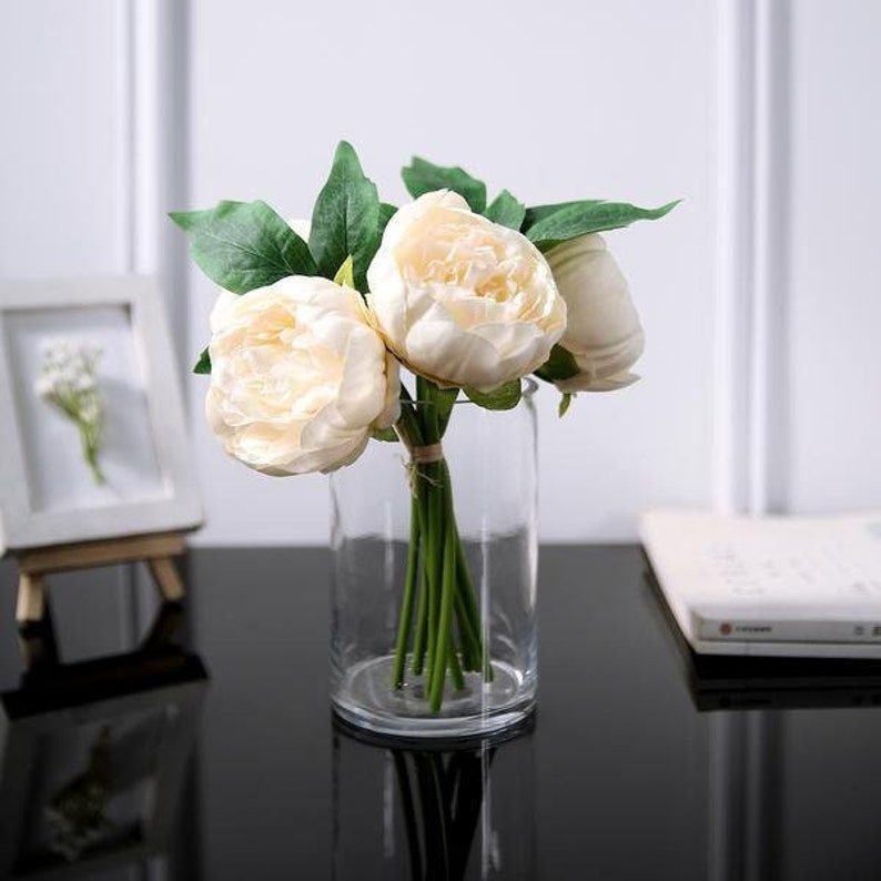 Pu&ograve; includere: Un vaso di vetro trasparente contiene un bouquet di fiori di peonia color avorio con foglie verdi. I fiori sono fitti e gli steli sono legati con spago. Una piccola cornice e un libro sono sullo sfondo.