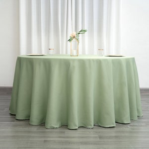 Op de afbeelding: Een ronde, saliegroene tafelkleed met een gladde, kreukvrije afwerking. Het tafelkleed ligt op een houten vloer en heeft een lichte drape aan de randen.