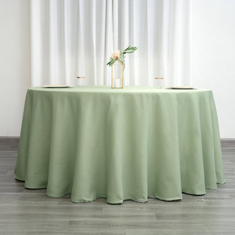 Op de afbeelding: Een ronde, saliegroene tafelkleed met een gladde, kreukvrije afwerking. Het tafelkleed ligt op een houten vloer en heeft een lichte drape aan de randen.