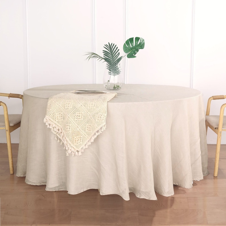 May include: A round, beige tablecloth with a crocheted runner in the center. The tablecloth is draped over a round table and has a subtle sheen.