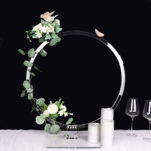 May include: A clear acrylic wedding arch with a circular design, adorned with white flowers, green foliage, and two gold butterfly accents. The arch is set on a clear base, with white candles and wine glasses in the foreground.