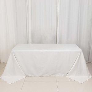 May include: A white rectangular tablecloth draped over a table. The fabric is smooth and appears to be made of a lightweight material. The tablecloth covers the entire table surface, with the corners hanging down.
