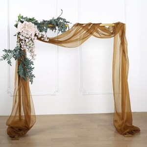 May include: A gold metal archway decorated with a sheer brown fabric drape and a garland of white roses and green leaves.