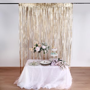 May include: Gold foil fringe curtain backdrop for a party or event. The curtain is hung from a black rod and is in front of a white wall. A white table with a white tablecloth is in front of the curtain. There are flowers and a cake stand on the table.