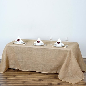 May include: A rustic brown burlap tablecloth with a natural linen texture. Three white dinner plates with white napkins folded into a rose shape and a single red rose on top of each napkin.  The plates are set on a wooden table with two wine glasses on each side.