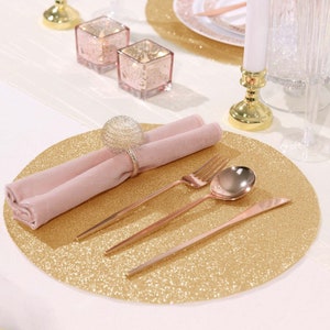 May include: Gold glitter charger plate with a pink napkin, a silver napkin ring, a fork, a knife, and a spoon. The charger plate is on a white table.