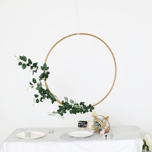 May include: A gold metal hoop with greenery and white flowers draped around it. The hoop is hanging from the ceiling above a table with a white tablecloth. There is a small chalkboard sign on the table that says "Welcome to the party".