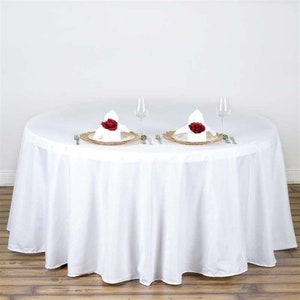 May include: White round tablecloth with a 108-inch diameter, set for a formal dinner with two place settings. Each place setting includes a gold charger plate, white napkin with a red rose, and silverware.