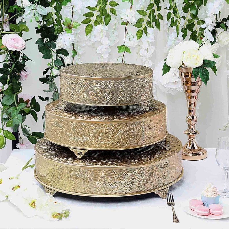 May include: Three gold metal cake stands with an ornate floral design. The stands are stacked on top of each other, with the largest stand on the bottom and the smallest stand on top. The stands have a round shape and are decorated with a raised floral pattern.