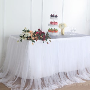 May include: A white rectangular table covered with a white tulle skirt. The table is set with a three-tiered cupcake stand holding cupcakes, a bouquet of flowers, and two wine glasses. A floral arrangement of roses is on the table.
