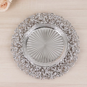 May include: A silver charger plate with an ornate, scrolling floral design around the rim. The center of the plate has a radial pattern. The plate is made of a shiny, reflective material, and is suitable for special occasions.