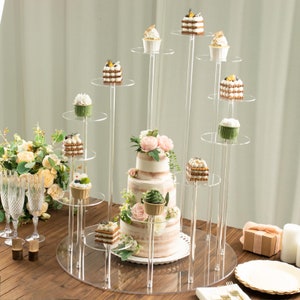 May include: A tiered acrylic cake stand with a white cake decorated with pink flowers and green leaves. The stand has multiple levels for displaying cupcakes, pastries, and other desserts.