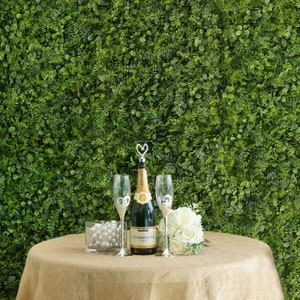 May include: A champagne bottle with a heart-shaped stopper and two champagne flutes on a burlap tablecloth in front of a green artificial wall.