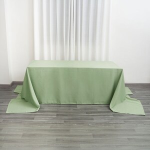 May include: A sage green rectangular tablecloth draped over a table. The fabric hangs over the edges, with the corners touching the floor. The background includes a white curtain and a gray wooden floor.