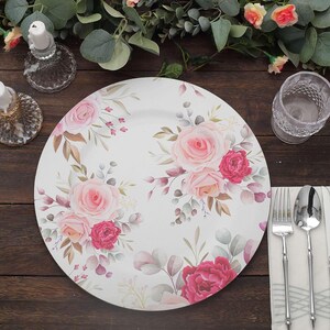 May include: White ceramic dinner plate with a floral design featuring pink and red roses, green leaves, and brown accents. The plate is surrounded by silverware, a glass, and decorative greenery. A beautiful addition to any table setting.