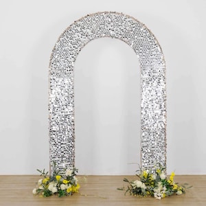 May include: A large, silver sequin-covered archway, ideal for weddings or events. The arch is decorated with small, reflective sequins, creating a shimmering effect. The base of the arch is adorned with arrangements of white and yellow flowers, adding a touch of natural beauty.