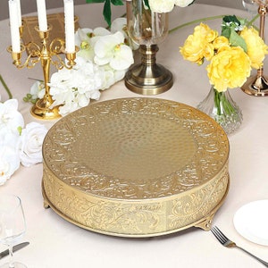 May include: Gold metal cake stand with an ornate, embossed design. The stand has three legs and a round top.