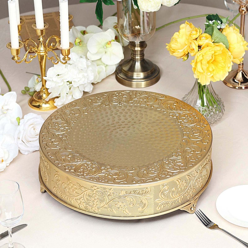 May include: Gold metal cake stand with an ornate, embossed design. The stand has three legs and a round top.