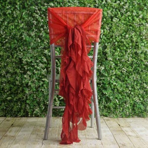 May include: A red chair cover with a ruffled design is draped over a silver chair. The chair cover is made of a sheer fabric and is tied in a bow at the top. The background is a green wall.