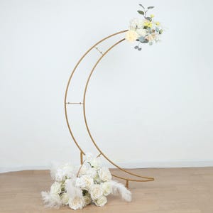 May include: A gold metal crescent moon arch with white roses and feathers at the base and a bouquet of white and yellow flowers at the top. The arch is a decorative piece for weddings or events.