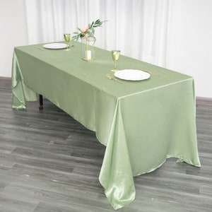 May include: A sage green satin tablecloth draped over a rectangular table with two place settings and a vase of flowers in the center.