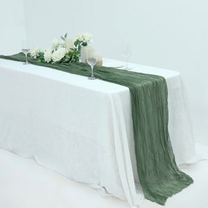 May include: A white tablecloth with a green linen table runner. The table runner is draped over the table and has a textured, crinkled look. There are two glasses and a small bouquet of white flowers on the table.