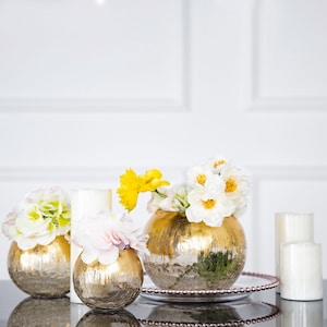 May include: Three gold metallic vases with different floral arrangements. The vases are round and have a hammered texture. The flowers are white and yellow.