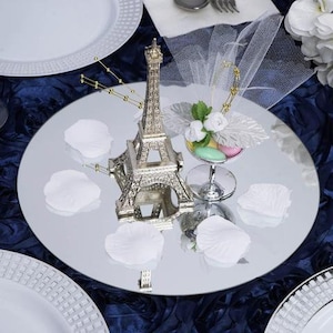 May include: A silver mirrored round charger plate with a silver Eiffel Tower decoration, white flower petals, and a small glass bowl with candy on top.