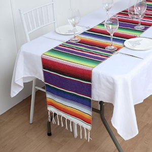 May include: A colorful Mexican-style table runner with a fringe border. The runner is made of woven fabric and features a traditional striped pattern in shades of red, yellow, green, blue, and purple.