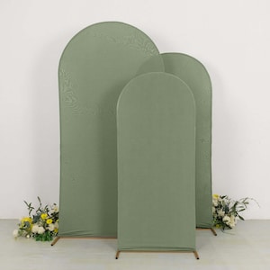 May include: Three sage green arch-shaped backdrops with a textured fabric. The backdrops are of varying heights and widths, with a simple gold-colored metal frame at the base. Floral arrangements with white and yellow flowers are placed at the base of the backdrops.