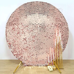 May include: A large, round backdrop covered in shimmering rose gold sequins. The backdrop is supported by a gold metal stand. Tall, gold candle holders with lit candles and small gift boxes are in front of the backdrop.