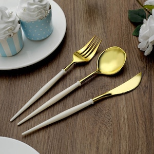 May include: A gold-colored fork, spoon, and knife set with white handles. The silverware is arranged on a wooden surface, next to cupcakes in blue and white patterned wrappers. The utensils are for dining.
