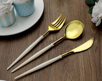 May include: A gold-colored fork, spoon, and knife set with white handles. The silverware is arranged on a wooden surface, next to cupcakes in blue and white patterned wrappers. The utensils are for dining.