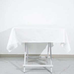 May include: A white tablecloth draped over a white folding table. The table has a square top and is set against a white wall. The tablecloth hangs over the edges of the table.