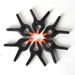 May include: Six black plastic clamps with orange handles arranged in a circle. The clamps are used for holding objects together.