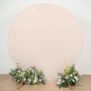 May include: A large, round, blush pink backdrop with two floral arrangements at the base. The arrangements are made up of white flowers, green leaves, and yellow flowers.