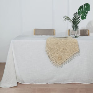 May include: A white linen tablecloth with a crocheted beige table runner with fringe. The table runner has a geometric pattern and is placed on a white table with two chairs.