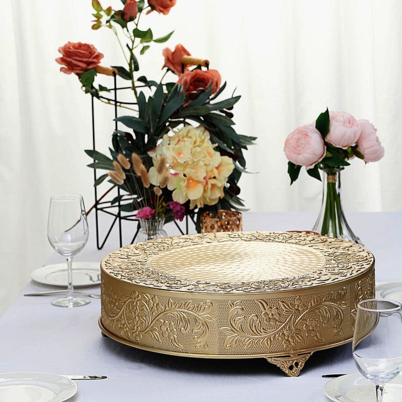 May include: Gold metal cake stand with an ornate design. The stand has a round top and three legs.