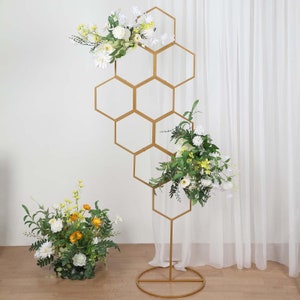 May include: A gold metal hexagonal archway with a geometric design. The archway is decorated with white and yellow flowers. The archway is standing on a white surface.