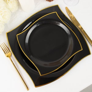 May include: Black square plates with gold trim, stacked on a white surface. A gold fork and knife are placed on the side. The plates have a unique wavy edge design. The plates are likely for a formal event or dinner party.