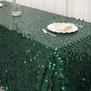 May include: A dark green sequin tablecloth covers a rectangular table. A white plate with a folded beige napkin and silverware are set on the table. A clear glass vase with a single flower is visible.