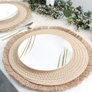 May include: A white plate with gold brushstroke accents sits atop a woven beige placemat with a fringed edge. Gold cutlery rests beside the plate, and another plate is visible in the background. The setting suggests a dining or event theme.