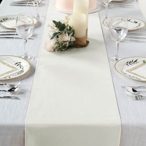 May include: A cream-colored table runner on a white tablecloth, set for a formal occasion. The table is decorated with candles, floral arrangements, plates with gold trim, silverware, and wine glasses. The runner adds a touch of elegance.