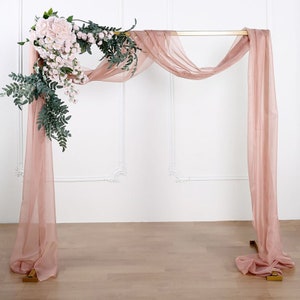 May include: A gold metal archway decorated with a blush pink sheer fabric backdrop and a floral garland of pink roses and greenery.